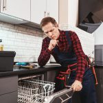 Solve the problem. Male technician think about fixing dishwasher in kitchen with instruments and smiling